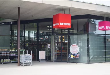 A modern supermarket with large glass sliding doors. Above the entrance, red signs hang with the names "M-Preis" and "BIPA".