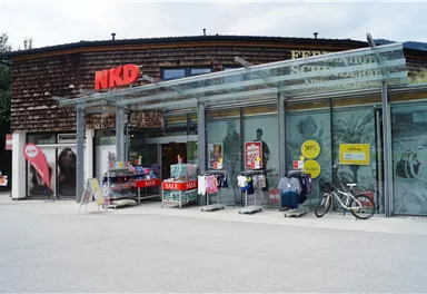 Ein Geschäft von NKD mit einer modernen Glasfassade. Vor dem Laden stehen Kleidung und Werbetafeln mit Angeboten.