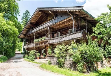 A traditional wooden house with several balconies, surrounded by green trees and plants. The path leads to the house, creating an inviting atmosphere.