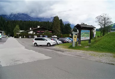 Ein Parkplatz in einer malerischen Umgebung mit Bergen im Hintergrund. Auf der linken Seite ist ein Informationsschild zu sehen.