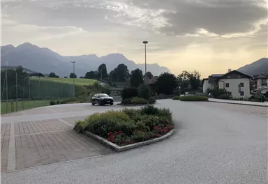 A peaceful landscape with mountains in the background and a road. Some flowerbeds and a parked car are also visible.