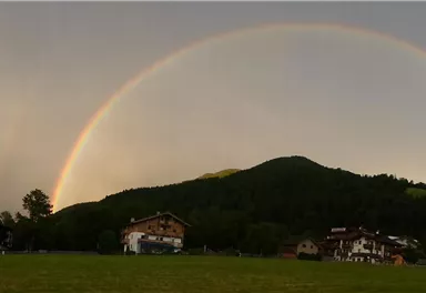 Ein Regenbogen überspannt die Landschaft mit sanften Hügeln. Im Vordergrund sind einige Häuser und grüne Wiesen zu sehen.