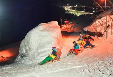 Ein nächtliches Winter-Szenario mit mehreren Personen, die auf Schlitten den schneebedeckten Hang hinunterfahren. Die Umgebung ist beleuchtet und es gibt einen großen Schneehaufen in der Nähe.