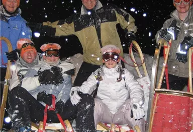 Eine Gruppe von sechs Personen posiert im Schnee mit Schlitten. Alle tragen Helme und Sonnenbrillen, während es schneit.