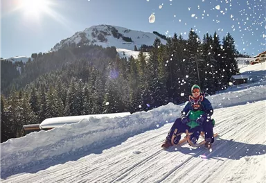 Zwei Personen fahren auf einem Schlitten den verschneiten Hang hinunter. Im Hintergrund sind Bäume und Berge zu sehen, während die Sonne strahlt.