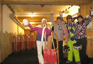 Eine Gruppe von vier Menschen in Winterkleidung steht in einem Skiraum. Sie lachen und halten ihre Helme sowie Schlitten in den Händen.