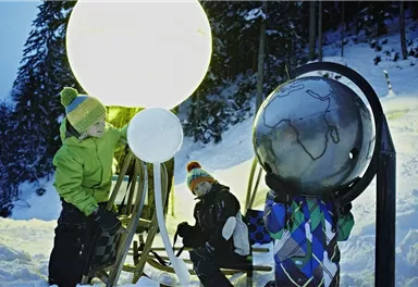 Zwei Kinder spielen im Schnee mit großen, leuchtenden Objekten. Im Hintergrund sind Bäume und eine winterliche Landschaft zu sehen.