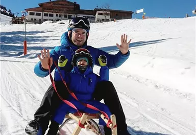 Ein Vater und sein Kind sitzen zusammen auf einem Schlitten im Schnee. Im Hintergrund sieht man eine Berghütte und strahlend blauen Himmel.