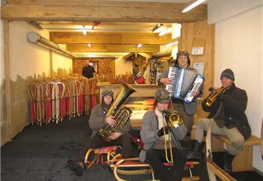 Eine Gruppe von Musikern spielt Instrumente in einem Raum mit Schlitten im Hintergrund. Sie tragen winterliche Kleidung und scheinen Spaß zu haben.