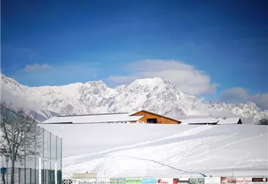 A snow-covered landscape with majestic mountains in the background. A wooden building stands in the foreground, under a clear blue sky.