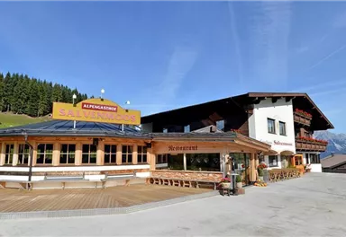 A picturesque restaurant in the mountains with a large terrace.  
Surrounded by green meadows and fir forests under a clear blue sky.