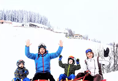 Eine Familie sitzt auf Schlitten im Schnee und lächelt fröhlich. Im Hintergrund sind verschneite Bäume und Hütten zu sehen.