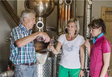 An older man is serving drinks to two women in a distillery. The women are watching with interest and holding glasses in their hands.