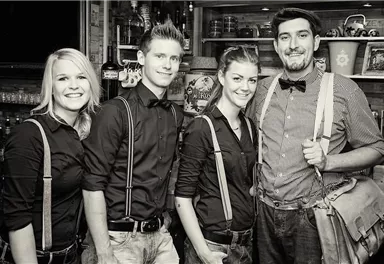 A group of four people stands in a bar, all wearing black and suspenders. They are smiling kindly at the camera.