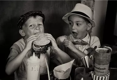 Two boys are sitting at a table. The boy on the left is biting into a large burger, while the other is watching in surprise.