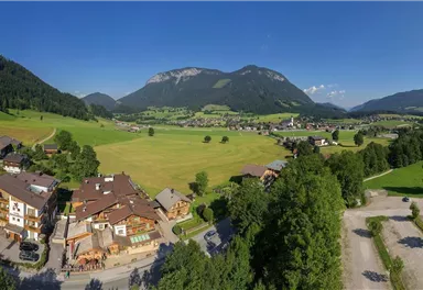 Eine malerische Landschaft mit grünen Wiesen, Bergen im Hintergrund und einem kleinen Dorf. Der Himmel ist klar und sonnig.