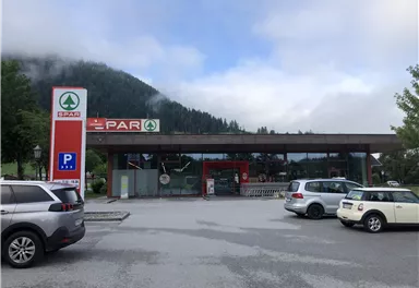 A SPAR store in a rural setting, surrounded by mountains and trees. In front of the shop is a parking lot with several cars.