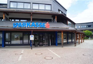 Eine moderne Bankgebäude mit dem Schriftzug „Sparkasse“ in blauer Schrift. Der Außenbereich ist gepflastert und es gibt einige Parkplätze.