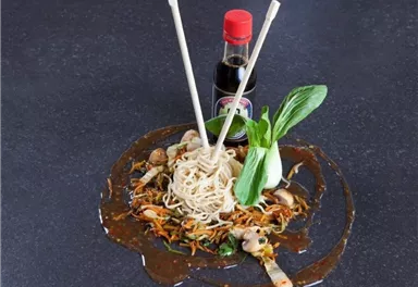 A bowl of Asian noodles surrounded by fresh vegetables. A small bottle of soy sauce and chopsticks complement the dish.