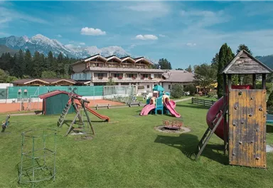 Ein kinderfreundlicher Spielplatz mit Rutschen und Schaukeln in einem grünen Garten. Im Hintergrund sieht man ein mehrstöckiges Gebäude und die Berge.