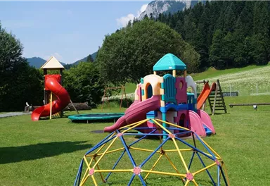 Ein Spielplatz mit bunten Spielgeräten und Rutschen. Im Hintergrund sind grüne Wiesen und Berge zu sehen.
