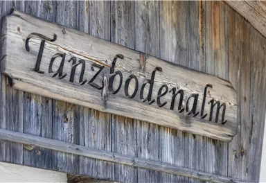 Ein Holzschild mit der Aufschrift „Tanzbodenalm“. Es befindet sich an einer rustikalen Wand.