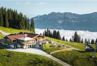 Eine malerische Berghütte umgeben von grünen Wiesen und hohen Bäumen. Im Hintergrund erheben sich majestätische Berge über einem Nebelmeer.