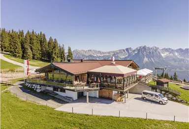 Eine alpine Berghütte mit schöner Aussicht auf die Berge. Umgeben von Bäumen und grünem Gras, bietet sie eine einladende Atmosphäre.