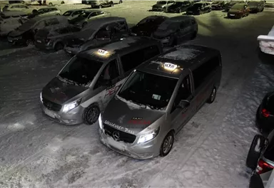 Two silver taxis are parked in a snowy parking lot. The vehicles are partially covered with snow, and more cars are visible in the background.