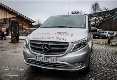 A silver taxi with the inscription "Sölleuken Pass" is parked on a cobblestone street. In the background, there is a wooden building and a rural setting.