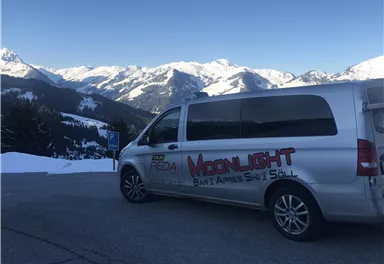 A silver van is parked in a parking lot with snow-covered mountains in the background. The surroundings are wintery and sunny.