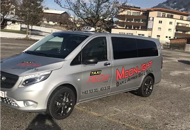 A silver taxi with the inscription "Moonlight" is parked in a parking lot. In the background, trees and buildings can be seen.