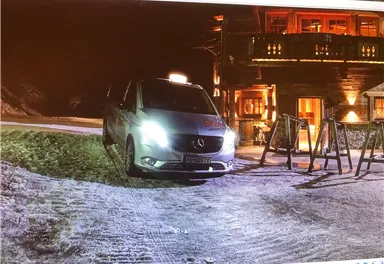 A Mercedes vehicle is parked on a snow-covered road at night. In the background, a lit building can be seen.