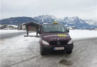 Ein Taxi steht auf einer schneebedeckten Straße mit einer malerischen Berglandschaft im Hintergrund. Die Umgebung ist winterlich und ruhig.