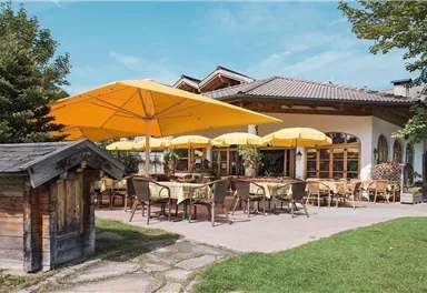 Ein einladendes Restaurant mit gelben Sonnenschirmen und einer gemütlichen Terrasse. Die umliegende Grünfläche und die Bäume schaffen eine angenehme Atmosphäre.