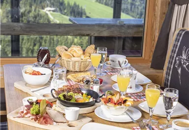Ein reichhaltiges Frühstücksbuffet mit verschiedenen Speisen und Getränken auf einem Tisch. Im Hintergrund sind die grünen Berge und ein klarer Himmel zu sehen.