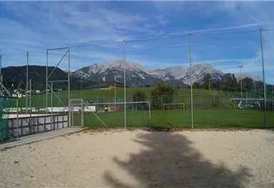 A sand court in front of a sports facility with a view of majestic mountains. The sky is clear and the surroundings are green and inviting.