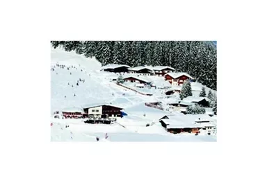 A winter landscape with snow-covered hills and several wooden houses. In the background, trees can be seen, and some people are skiing.