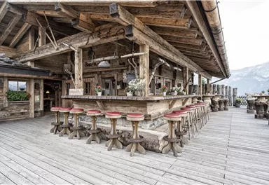 A cozy mountain cabin with a wooden bar and several stools. Mountains can be seen in the background.