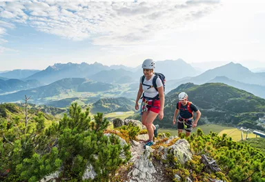 Zwei Kletterer wandern auf einem Berggipfel mit Blick auf die umliegenden Berge und das grüne Tal. Die Landschaft ist von einer klaren, blauen Himmel umgeben.