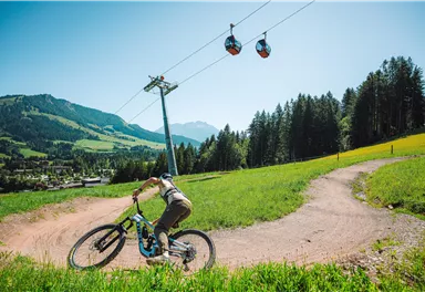 Ein Mountainbiker fährt auf einem kurvigen Pfad durch eine grüne Wiese. In der Nähe fahren Gondeln über die Landschaft.