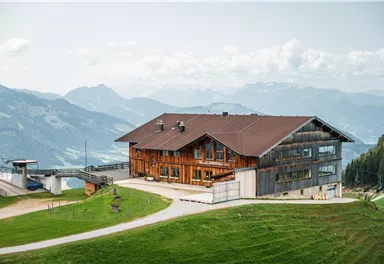 Ein rustikales Holzhaus in einer malerischen Berglandschaft. Im Hintergrund sind beeindruckende Berge und ein klarer Himmel zu sehen.