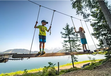 Zwei Kinder spielen auf einer Wackelbrücke am Wasser. Im Hintergrund sind Berge und Bäume sichtbar.