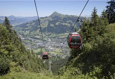 A cable car travels over a green landscape with a view of the mountains. Below, a picturesque valley stretches out with small houses and plenty of nature.