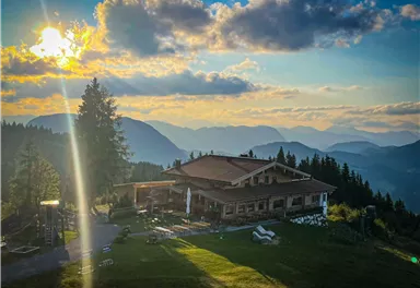 Eine charmante Berghütte umgeben von majestätischen Bergen. Die Sonne geht hinter den Gipfeln unter und taucht die Landschaft in warmes Licht.