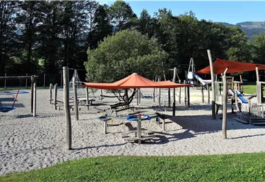 Ein Spielplatz mit Sandboden und verschiedenen Spielgeräten. Schattensegmente bieten Schutz vor der Sonne.