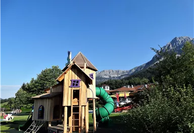 Ein Spielplatz mit einem Holzspielhaus und einer grünen Rutsche. Im Hintergrund sind Berge und Bäume zu sehen.