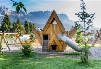 A creative playground with a wooden playhouse and a slide. In the background, green trees and impressive mountains can be seen.