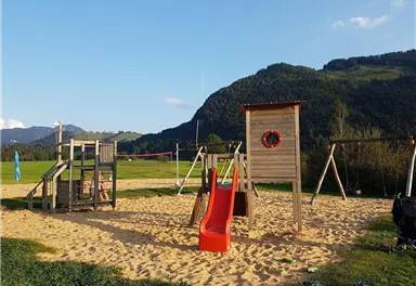 Ein Spielplatz mit einer Rutsche und Schaukeln auf sandigem Boden. Im Hintergrund sind grüne Wiesen und Berge zu sehen.