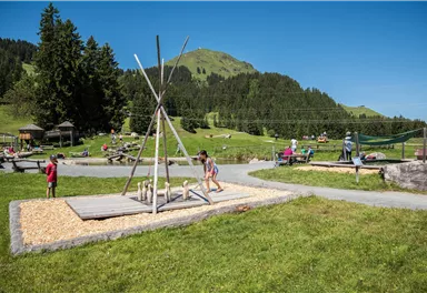 A playground in a beautiful, green landscape with hills in the background. Children are enjoying the various play opportunities under a clear blue sky.